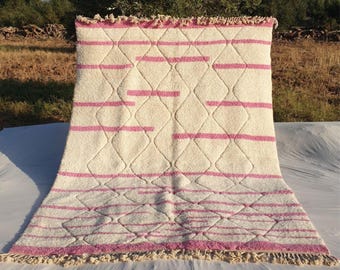 May include: A white and pink Moroccan rug with a diamond pattern and pink stripes. The rug has a fringe border.