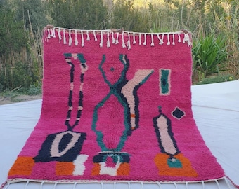 May include: A pink Moroccan rug with a geometric design in black, white, green, and orange. The rug has a fringe border.