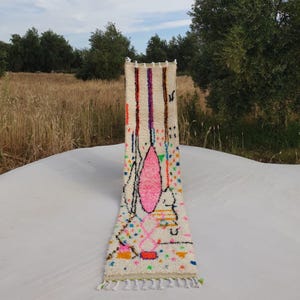 Könnte beinhalten: Ein weißer, handgewebter Teppich mit einem farbenfrohen, abstrakten Design. Der Teppich hat ein pinkfarbenes, orangefarbenes, gelbes, grünes, blaues und violettes Design. Der Teppich hat Fransen am unteren Ende.