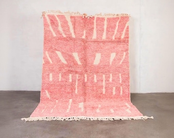 May include: A rectangular, hand-woven rug with a pink background and cream-colored geometric patterns. The rug has a thick pile and a fringe along the bottom edge. The rug is displayed against a white wall and a gray floor.