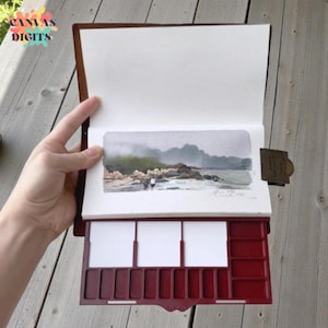 Op de afbeelding: Een bruin leren schetsboek met een aquarel van een kustlandschap. Het schetsboek heeft een ingebouwd rood palet met witte verfputten. De schildering toont een strand met figuren, rotsen en een mistige achtergrond. De tekst "CANVAS DIGITS" staat linksboven.