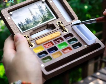 May include: A wooden watercolor paint set with a landscape painting and a variety of colorful paint blocks. The set includes a small white water container and a brush. The text "CANVAS DIGITS" is visible in the upper left corner.