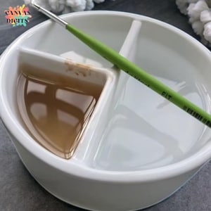 Puede incluir: Una paleta de pintura de cerámica blanca con tres secciones, una de ellas llena de pintura marrón. Un pincel verde descansa sobre la paleta. El texto "Canvas Digits" está en la esquina superior izquierda.