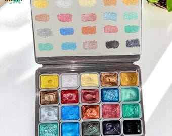 May include: A watercolor paint set with 24 metallic colors in a clear plastic case. The set includes a color chart with swatches of each color. The colors range from gold and silver to various shades of blue, green, and red.