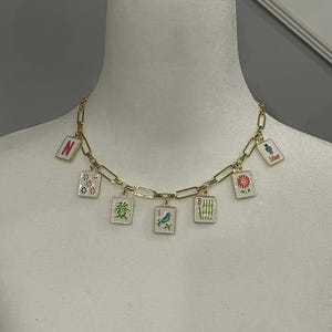 May include: A gold-toned chain necklace featuring seven rectangular charms, each decorated with colorful Mahjong tile designs including a bird, bamboo, flowers, and the letter N. The charms are evenly spaced along the polished metal link chain.