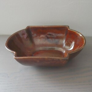 May include: A brown ceramic bowl with an unusual, asymmetrical form. The bowl has a glossy finish with reddish-brown tones. The rim has a slight lip, and the design is modern and artistic. The bowl is likely to be around 20 cm in diameter.