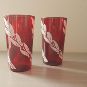May include: Two red drinking glasses with a white leaf design. The glasses are tall and cylindrical, with the leaf pattern wrapping around the sides. The glasses are sitting on a light-colored surface.