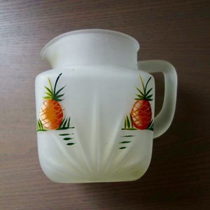 May include: White frosted glass pitcher with a handle. The pitcher has a pineapple design painted on the front with green leaves and orange fruit.