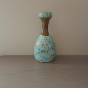 May include: A ceramic vase with a light blue body and a brown neck. The vase has a unique design of wavy, textured lines in a darker shade of blue. The top rim is a darker brown color. The vase is a decorative item.