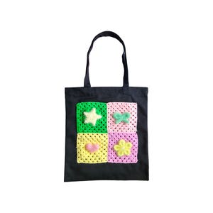 Puede incluir: Bolsa de tela negra con cuatro cuadrados de ganchillo que representan una estrella, una mariposa, un corazón y una flor. Los cuadrados son en tonos de verde, rosa, amarillo y blanco.