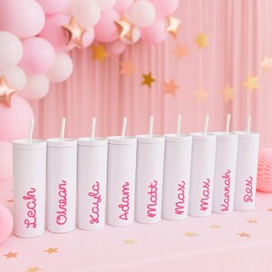 May include: White personalized tumblers with pink text and straws. The tumblers are on a pink surface, with pink balloons and gold star decorations in the background. Names on the tumblers include Leah, Orsan, Kayla, Adam, Matt, Max, Lannah, and Rex.