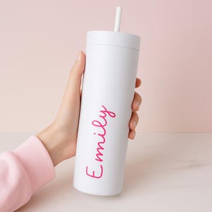 May include: A white tumbler with a matching lid and a white straw. The name "Emily" is written in pink script on the side. The tumbler is held against a soft pink background.