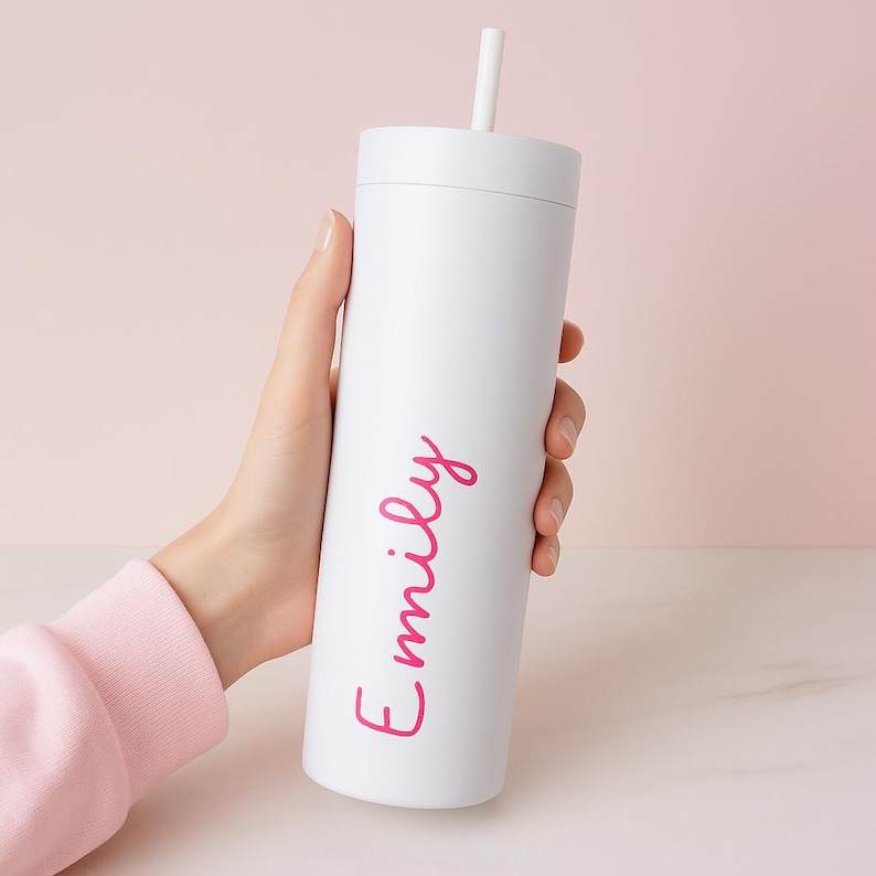 May include: A white tumbler with a matching lid and a white straw. The name "Emily" is written in pink script on the side. The tumbler is held against a soft pink background.
