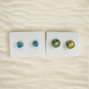 May include: Two pairs of stud earrings on white display cards. The earrings are round and have a blue and gold resin design. One pair features a gold seashell design.