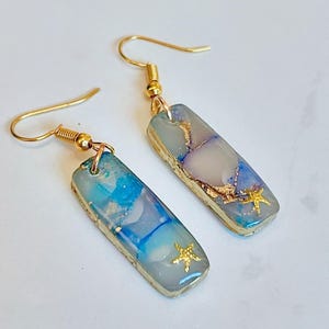 Peut inclure: Une paire de boucles d'oreilles pendantes dorées avec des accents en résine bleue et blanche et en feuille d'or. Chaque boucle d'oreille présente un petit charme d'étoile de mer dorée.