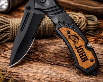 May include: Black folding pocket knife with a serrated blade and wooden handle. The handle features a fish graphic and the name "JOHN". The knife is shown on a wooden surface with rope and a flashlight in the background.