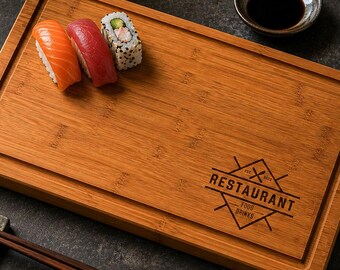 May include: A rectangular bamboo serving board with a grooved border and a laser-engraved logo reading RESTAURANT FOOD DRINKS. Three pieces of sushi, including salmon and tuna nigiri, sit on the board alongside dark wooden chopsticks on a rest.