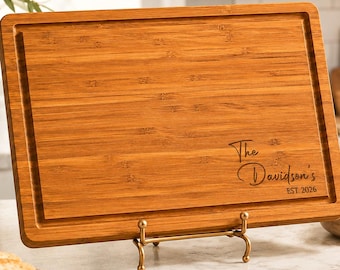 May include: A rectangular bamboo cutting board with a juice groove along the perimeter, resting on a gold metal stand. The board features laser-engraved text that reads The Davidson's EST. 2026 in a script and sans-serif font combination.