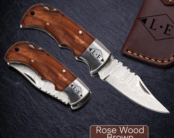 May include: Two folding knives with rosewood handles and patterned blades are displayed. The knives have silver accents and are next to a brown leather case with the initials "L.E." The text "Rose Wood Brown" is at the bottom.