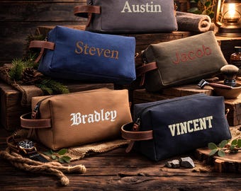 May include: Five personalized canvas toiletry bags in various colors: gray, blue, olive green, tan, and navy. Each bag features a name embroidered in a contrasting color, with brown leather handles and zipper pulls. The bags are displayed on a rustic wooden surface.
