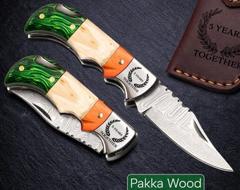 May include: Two folding knives with Pakka Wood handles. The handles feature green, cream, and orange sections. The blades have a patterned design, and the metal portion is engraved with "5 YEARS TOGETHER". A brown leather case is visible.
