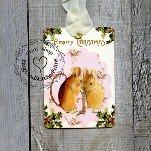 May include: A rectangular gift tag featuring a vintage illustration of two brown mice holding a small striped stocking. The tag has the text A merry CHRISTMAS at the top, a pink floral background, and holly leaf borders. A cream ribbon is attached to the top.