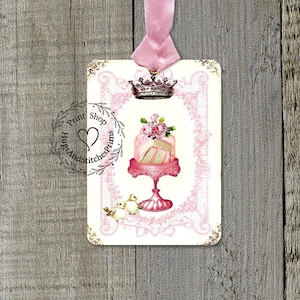 May include: Pink and white cake with a crown on top, surrounded by a pink floral design. The cake is on a pink pedestal with two white birds on the side. The image is in a vintage style frame with a pink background.