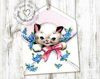 May include: A hanging tag featuring a white kitten with dark ears peeking out of a white envelope. The envelope is decorated with blue flowers and a pink bow. The text HugsAndStitchesPrims Print Shop is printed in a circular logo on the left side.