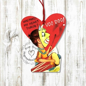 May include: A heart-shaped hanging ornament featuring a vintage illustration of a person wearing a yellow mask. The text reads, WHO MAKES MY HEART THROB? VOO DOO! A red string is attached to the top. The design includes a circular logo with the text Print Shop HugsAndStitchesPrims.