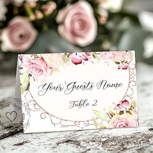 May include: A floral wedding place card with a watercolor design. The card features pink roses and green leaves, with the text "Your Guests Name" and "Table 2" in elegant script. A decorative border and logo are also present.