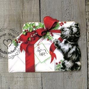 May include: A die-cut paper illustration featuring a white envelope wrapped in a large red ribbon with green holly leaves and red berries. A small light green gift tag with the handwritten text For me? is attached, and a black terrier dog stands beside it.