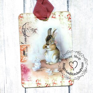 May include: A vintage-style postcard featuring a rabbit holding a teacup. The postcard has a floral border and a stamp that says "Posta Aerea". The image is printed on a wooden tag with a pink ribbon attached. The tag also has a stamp that says "Hugs And Stitches Prints Shop" with a heart in the center.