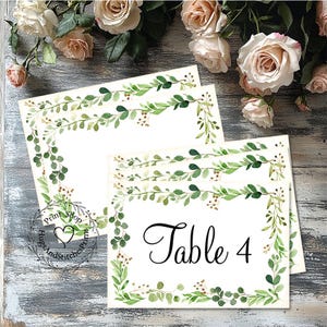 May include: White rectangular table cards with a green and brown botanical border. The cards have the text "Table 4" in black script. The cards are arranged on a rustic wooden surface with roses in the background.