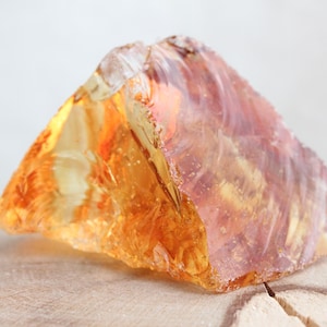 May include: A rough-cut citrine crystal, displaying a spectrum of orange and amber hues. The gemstone is presented on a light-coloured wooden surface, highlighting its natural form and colour variations.