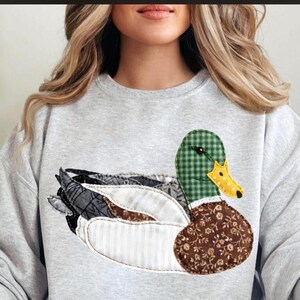 Peut inclure: Un sweat-shirt gris clair orné d'un appliqué de canard. Le canard présente une tête à carreaux verts, un bec jaune et une poitrine marron à motifs floraux. Les ailes sont faites de tissus gris, blancs et bruns.