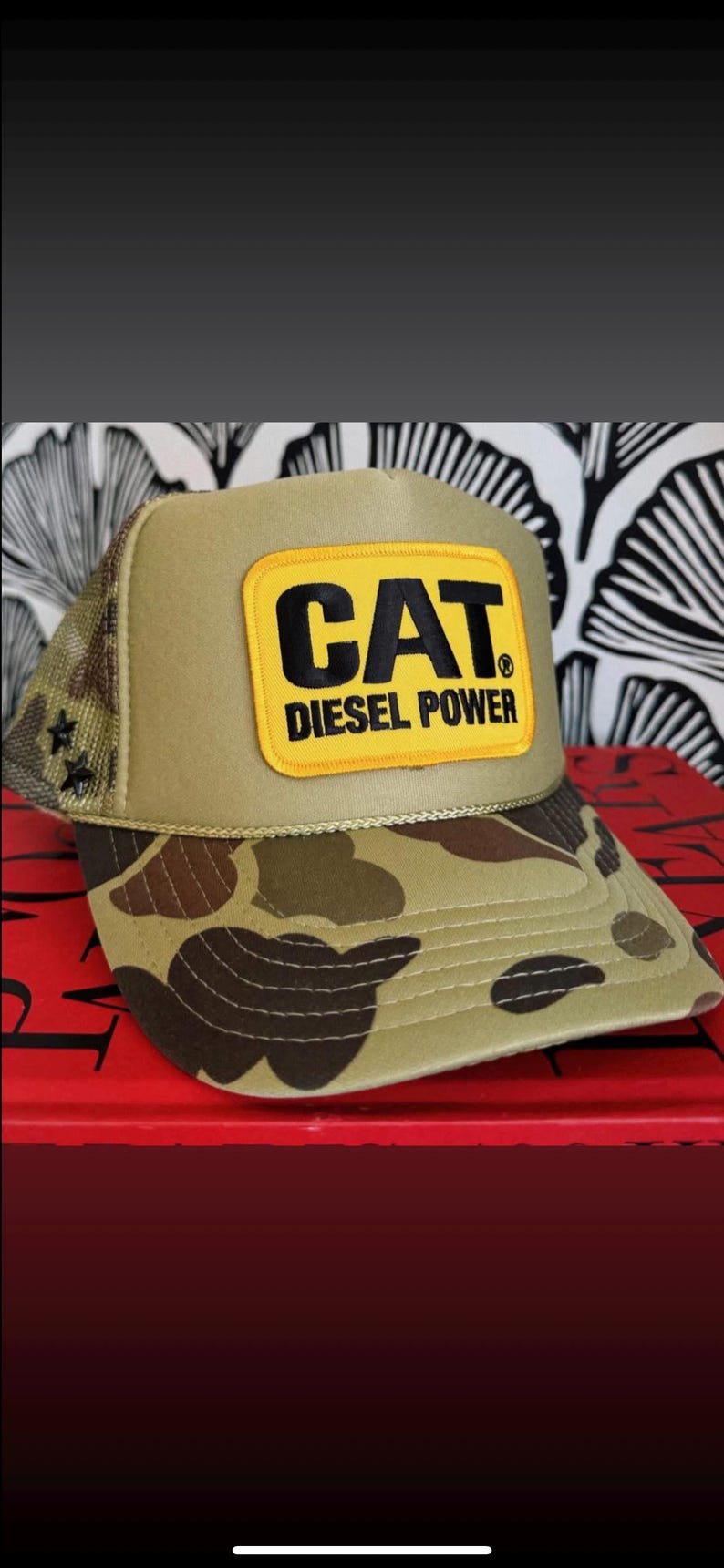 May include: A tan and camouflage trucker hat with a mesh back. The front features a yellow rectangle with the black text "CAT DIESEL POWER". Two black stars are on the left side of the mesh.