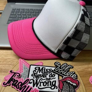 Op de afbeelding: Een truckerpet met een roze klep, witte voorkant en zwart-wit geblokte mesh achterkant. De pet ligt naast verschillende geborduurde patches met zinnen als "Trashy" en "Miss Never do no Wrong".