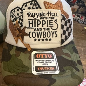 Op de afbeelding: Een witte trucker-pet met een camouflage-gaas achterkant. De voorkant van de pet heeft een witte patch met de tekst "Raising Hell with the Hippies and the Cowboys" en vijf sterren. De pet heeft ook verschillende patches, waaronder een ster, een geruit hart en een paar dobbelstenen.