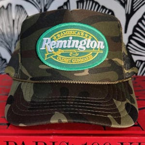 May include: Camouflage pattern trucker hat with a green and yellow oval patch that reads "America's Remington Oldest Gunmaker". The hat has a curved brim and a mesh back.