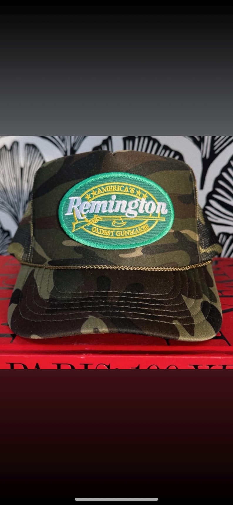 May include: Camouflage pattern trucker hat with a green and yellow oval patch that reads "America's Remington Oldest Gunmaker". The hat has a curved brim and a mesh back.