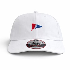 May include: White baseball cap with a red and blue flag logo embroidered on the front. The cap has a curved brim and a True Fit UPF 50+ label. A classic, casual accessory.