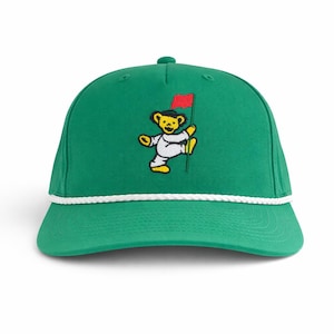 Può includere: Cappellino da baseball verde con un orsetto che gioca a golf. L'orso giallo indossa un completo bianco e tiene una bandiera rossa. Il cappellino ha un dettaglio in corda bianca e una visiera curva.