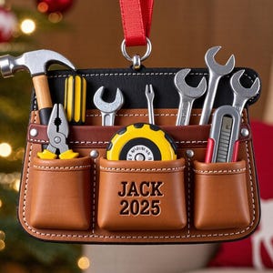 Könnte beinhalten: Ein 2D-flacher Weihnachtsschmuck in Form eines braunen Leder-Werkzeuggürtels mit Miniaturwerkzeugen wie Hammer, Schraubenschlüsseln und einem Maßband. Der Text "JACK 2025" ist sichtbar. Er hängt an einem roten Band.
