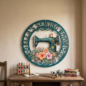 Peut inclure: Une enseigne murale circulaire bleu sarcelle avec le texte "ELLA'S SEWING ROOM" en police découpée. Le centre présente une illustration de machine à coudre Singer vintage entourée d'un arrangement floral coloré orange, rose et jaune.