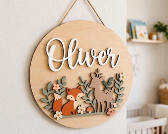 May include: A circular wooden wall sign featuring the name Oliver in white script lettering. The design includes layered wooden cutouts of a fox, a deer, small flowers, and foliage in shades of orange, green, and cream, suspended by a brown rope.