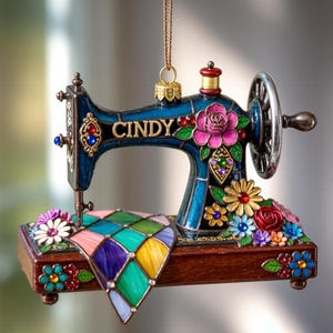 May include: A decorative sewing machine ornament in shades of blue, brown, and colourful floral accents. The word "CINDY" is displayed on the machine. The ornament features a quilt-like design and a gold-coloured hanging chain.