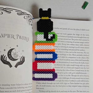 Peut inclure: Marque-page artisanal en perles de plastique colorées, en forme de pile de livres avec un chat noir au sommet. Le marque-page est inséré dans un livre ouvert sur le chapitre "Douze". Le marque-page mesure environ 18 cm de haut.
