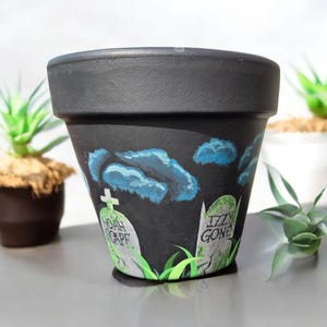 May include: Black terracotta planter with a spooky graveyard scene painted on it. The scene includes two tombstones with the names "Noah Gape" and "Izzy Gone" written on them. There are also green grass and blue clouds painted on the planter.