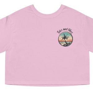May include: A pink cropped t-shirt featuring a graphic of a palm tree and the text "Tides and Vibes" in a circle on the left chest. The shirt has a Champion logo on the right sleeve.