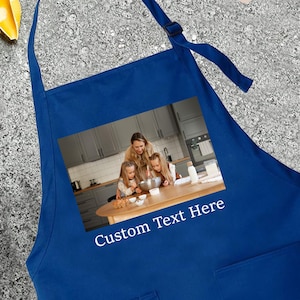 May include: A royal blue apron with a rectangular image of a family cooking in a kitchen. The apron has a neck loop and waist ties. The text "Custom Text Here" is printed on the apron.